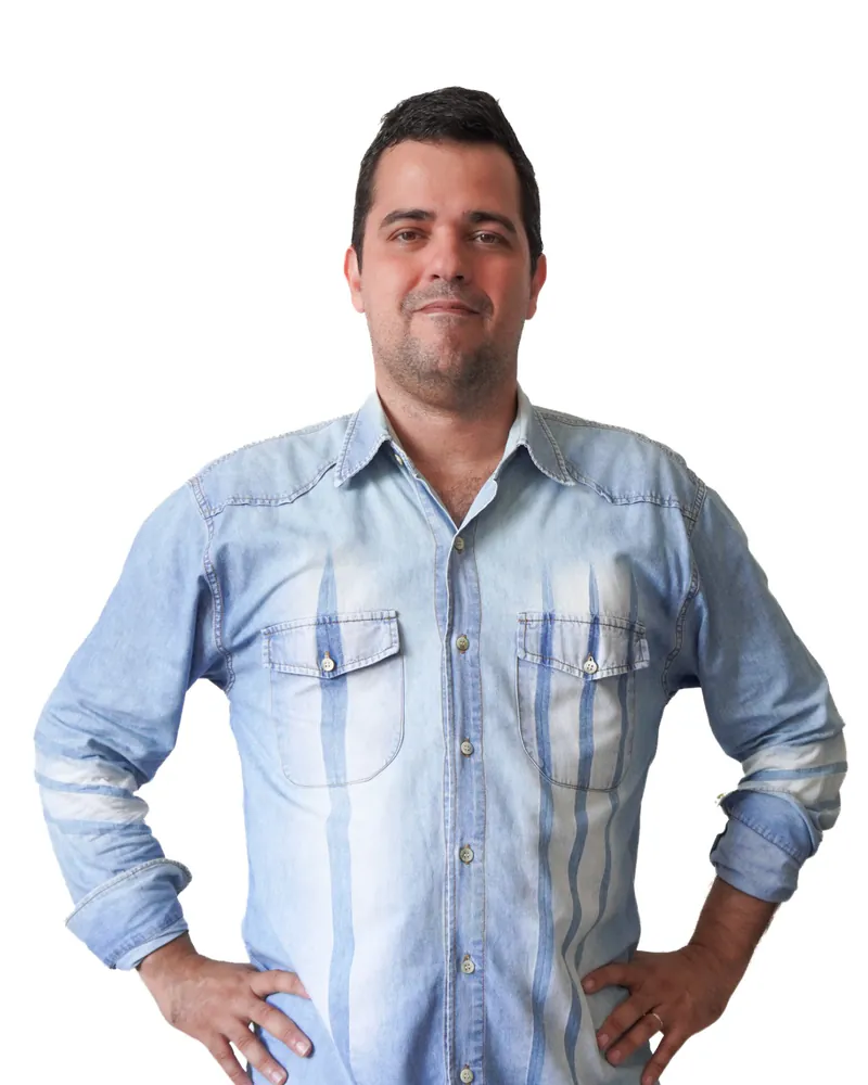 Homem de cabelo escuro e curto, com barba rala, sorrindo de leve, de camisa jeans azul-clara e com as mãos na cintura, diante de um fundo branco.